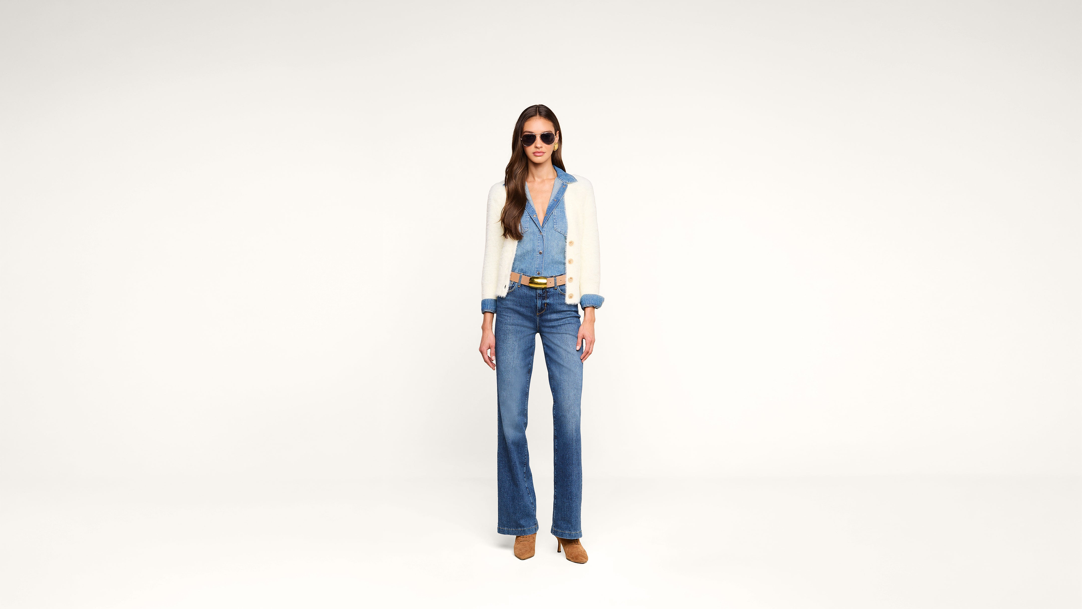 Woman wearing sunglasses, a light beige cardigan, a blue button-up shirt, blue high-waisted jeans, a brown belt, and brown shoes stands against a plain white background.