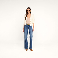 Una donna con gli occhiali da sole indossa l’Agathe Cardigan Top — un cardigan corto color crema dal tessuto soffice — abbinato a un top beige con scollo a V, jeans blu a vita alta, una cintura nera con fibbia dorata e scarpe marroni; il tutto su uno sfondo beige a tinta unita.