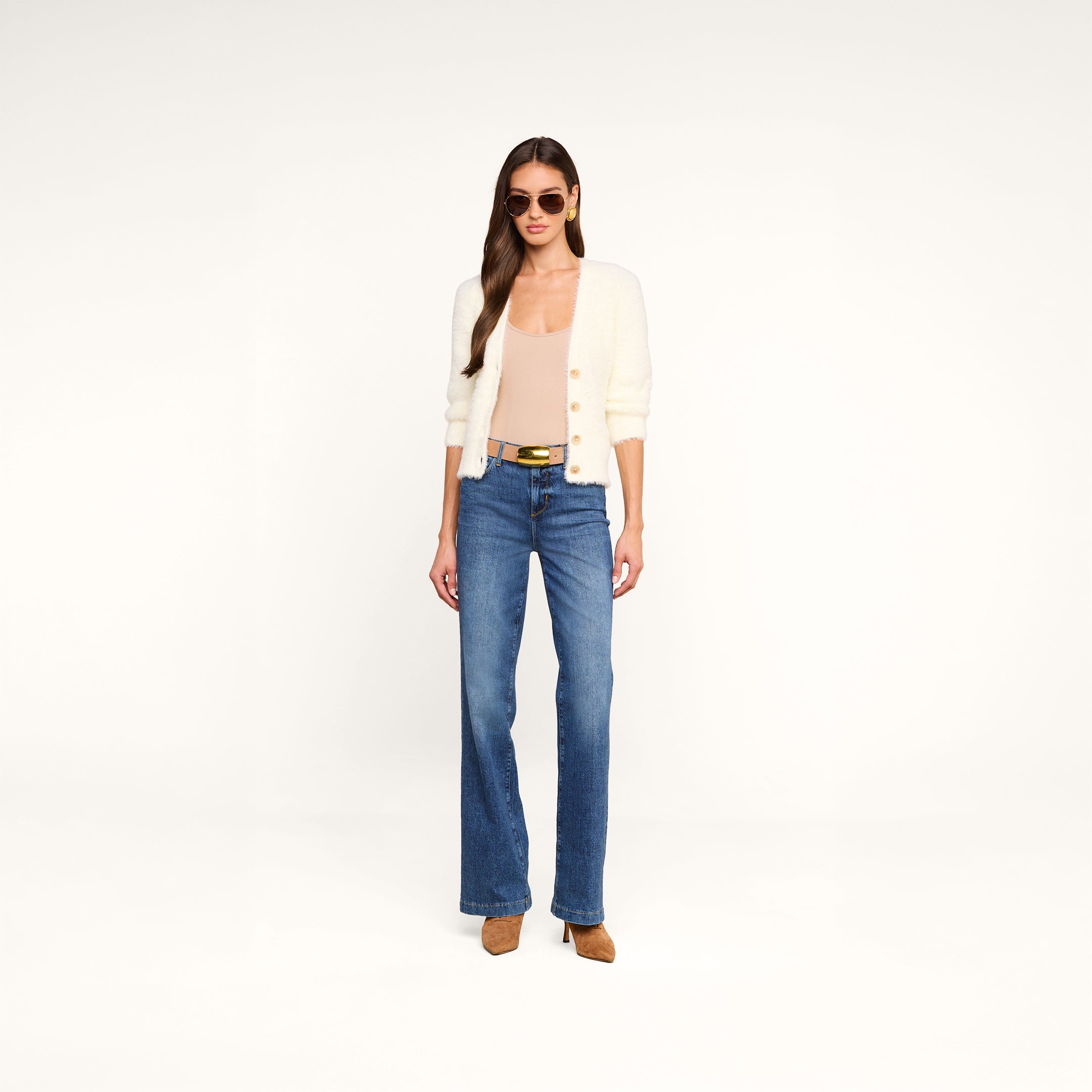 Una donna con gli occhiali da sole indossa l’Agathe Cardigan Top — un cardigan corto color crema dal tessuto soffice — abbinato a un top beige con scollo a V, jeans blu a vita alta, una cintura nera con fibbia dorata e scarpe marroni; il tutto su uno sfondo beige a tinta unita.