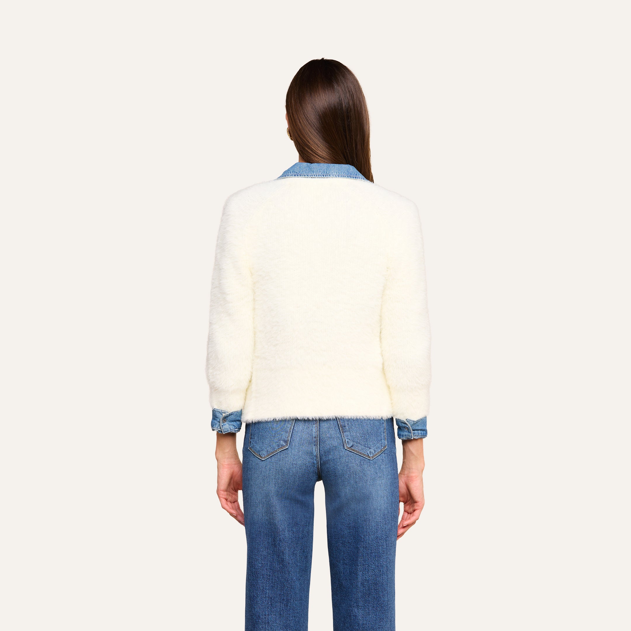 Una persona con lunghi capelli castani, che indossa il cardigan Agathe sopra una camicia di jeans blu e dei jeans blu, è ritratta di schiena su uno sfondo beige.