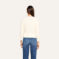 Si vede di spalle una persona con lunghi capelli castani che indossa l’Agathe Cardigan Top — un cardigan corto bianco e soffice — e dei jeans blu su uno sfondo beige.