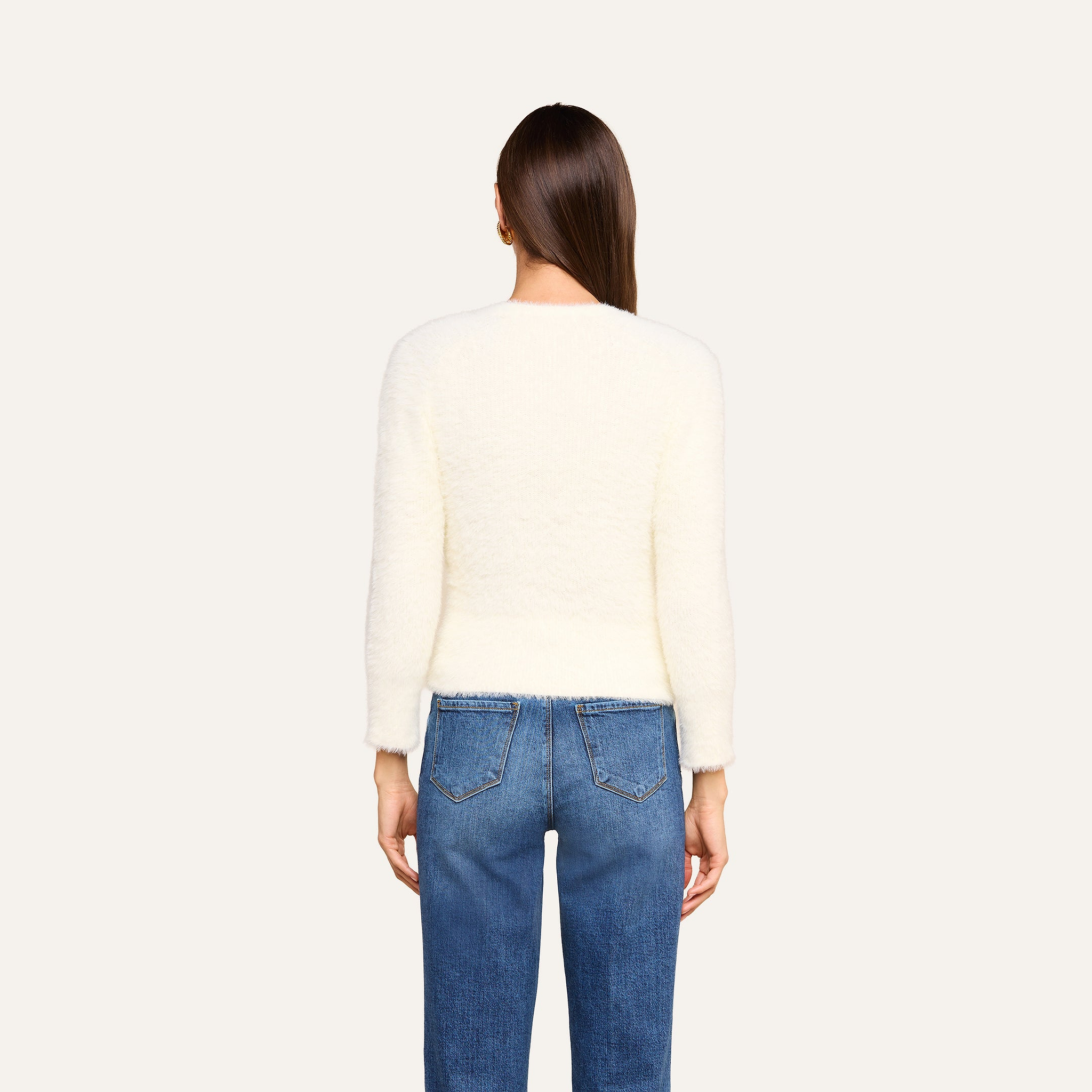 Si vede di spalle una persona con lunghi capelli castani che indossa l’Agathe Cardigan Top — un cardigan corto bianco e soffice — e dei jeans blu su uno sfondo beige.