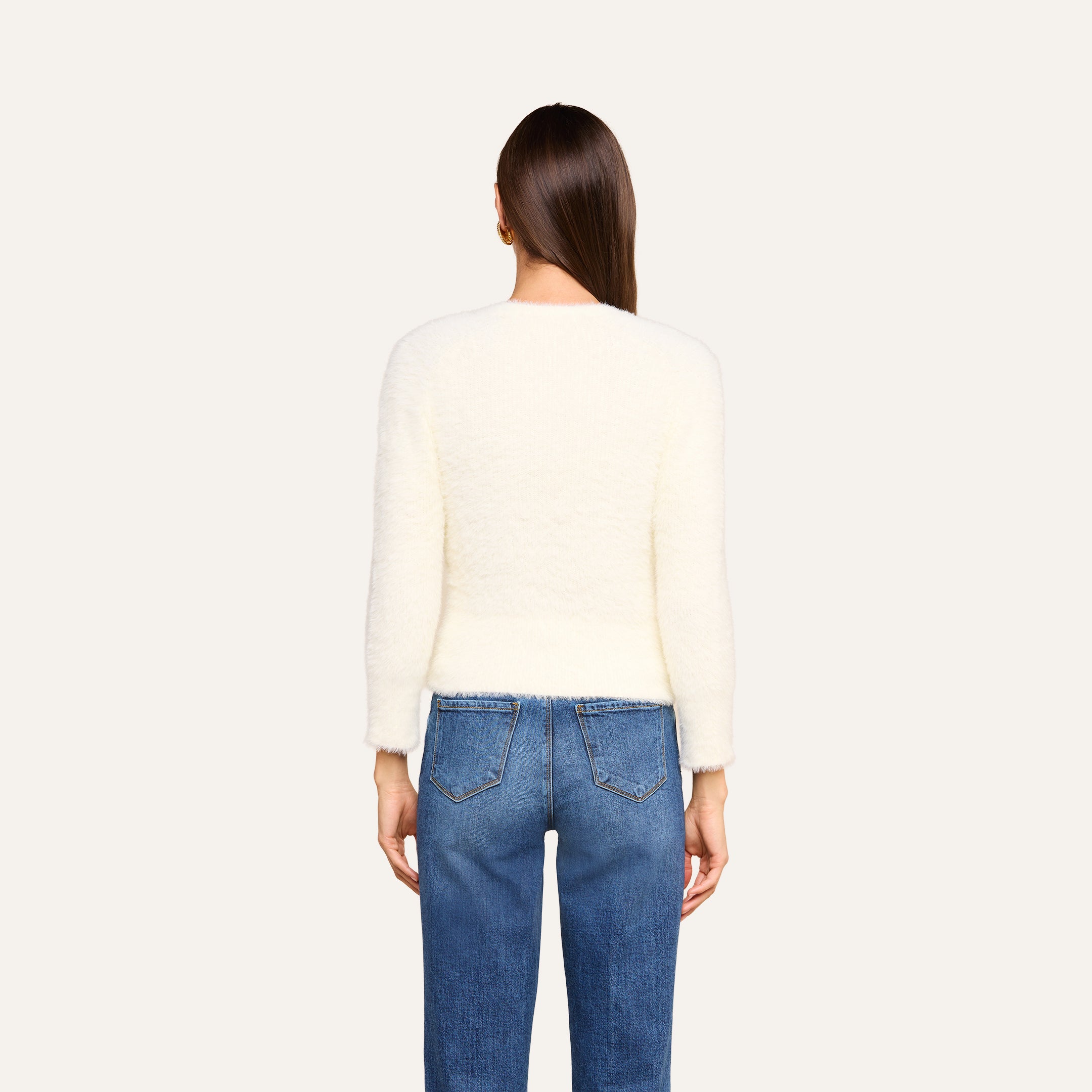 Si vede di spalle una persona con lunghi capelli castani che indossa l’Agathe Cardigan Top — un cardigan corto bianco e soffice — e dei jeans blu su uno sfondo beige.