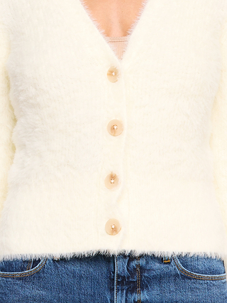 Une personne portant le cardigan « Agathe », un cardigan court blanc duveteux, superposé à un haut beige à col en V et associé à un jean bleu, photographiée des épaules aux hanches.