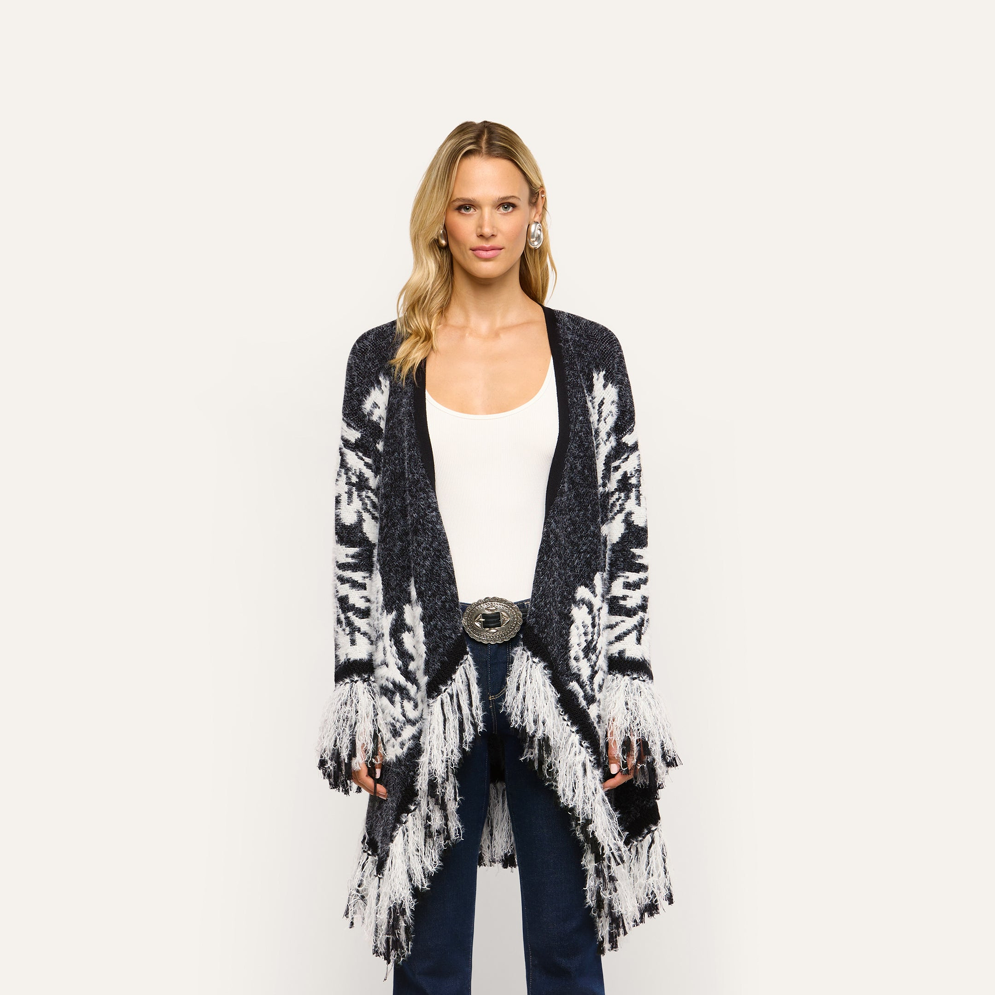 A person wearing a Naiad Jacquard Fringe Cardigan in a boho-chic, black and white pattern with long fringe trim along the hem and cuffs.