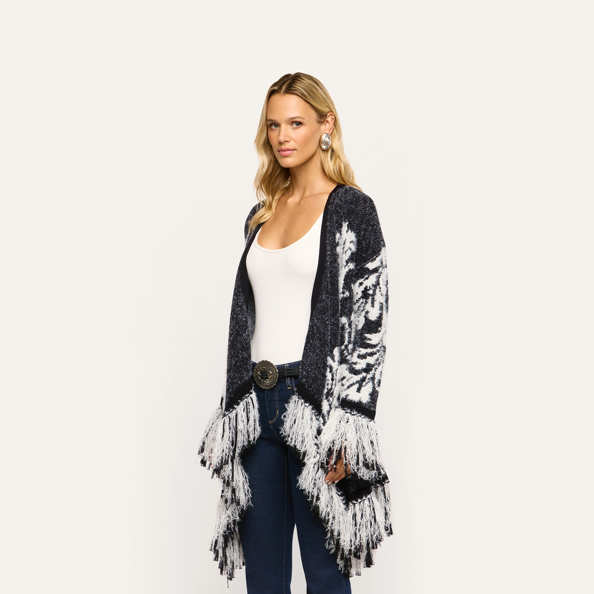 A woman wearing a Naiad Jacquard Fringe Cardigan in a boho-chic, black and white pattern with a relaxed fit and long fringe trim at the hem.