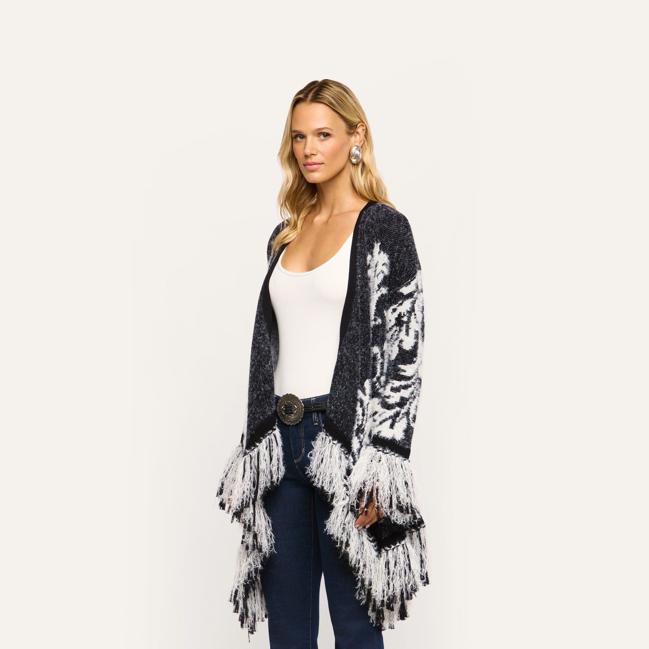 A woman wearing a Naiad Jacquard Fringe Cardigan in a boho-chic, black and white pattern with a relaxed fit and long fringe trim at the hem.