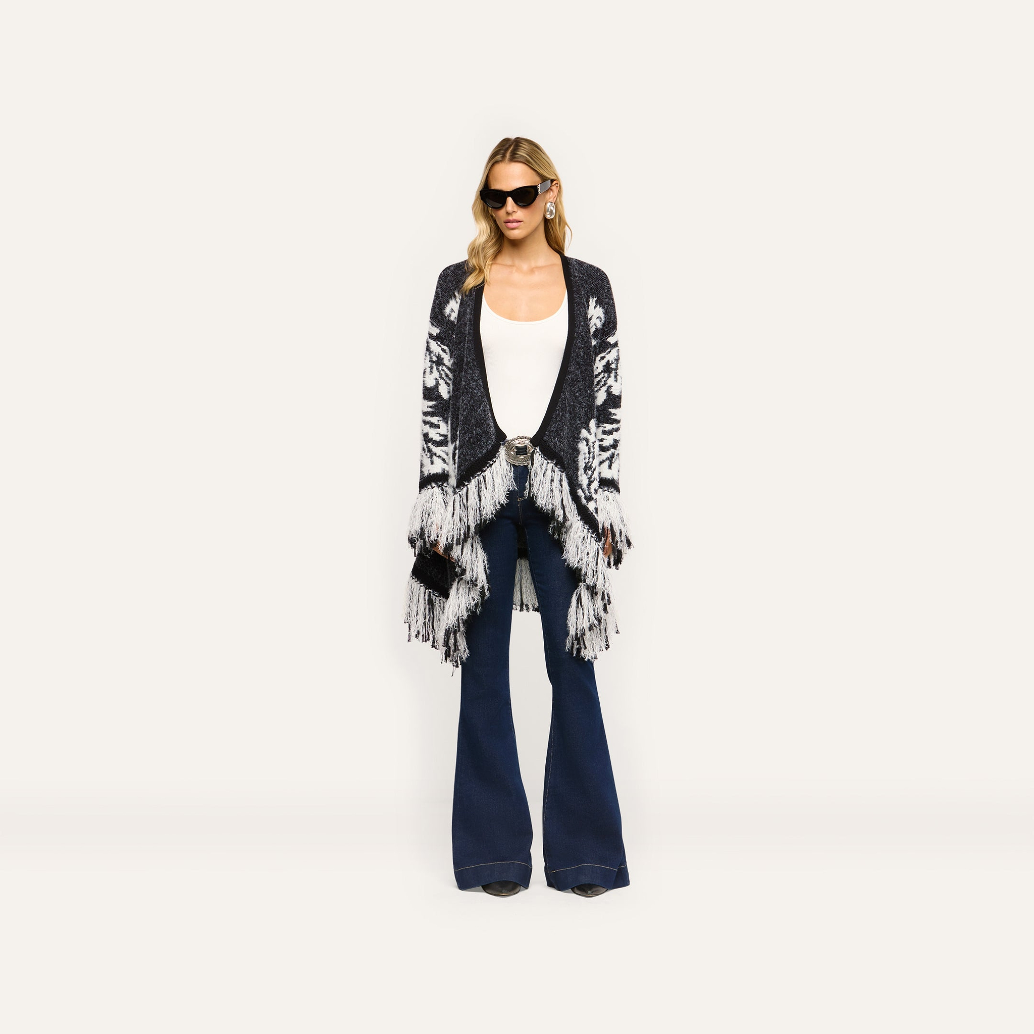 A person in a black and white patterned Naiad Jacquard Fringe Cardigan, white tank top, and relaxed-fit dark blue flared jeans.