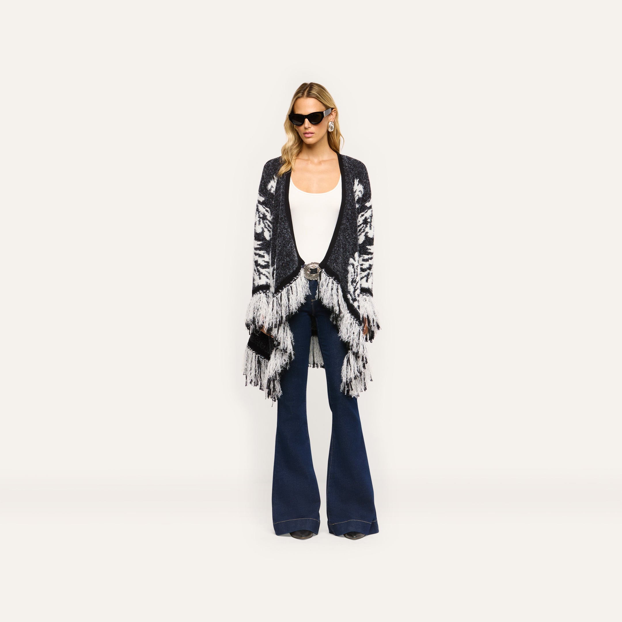 A person in a black and white patterned Naiad Jacquard Fringe Cardigan, white tank top, and relaxed-fit dark blue flared jeans.