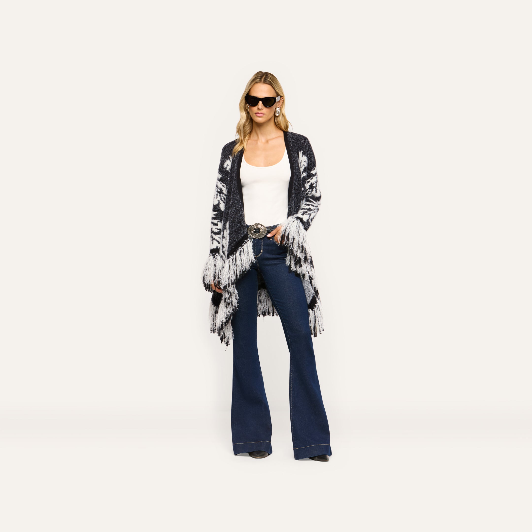 A woman wears a relaxed-fit, boho-chic black and white Naiad Jacquard Fringe Cardigan over a white tank top and flared blue jeans.
