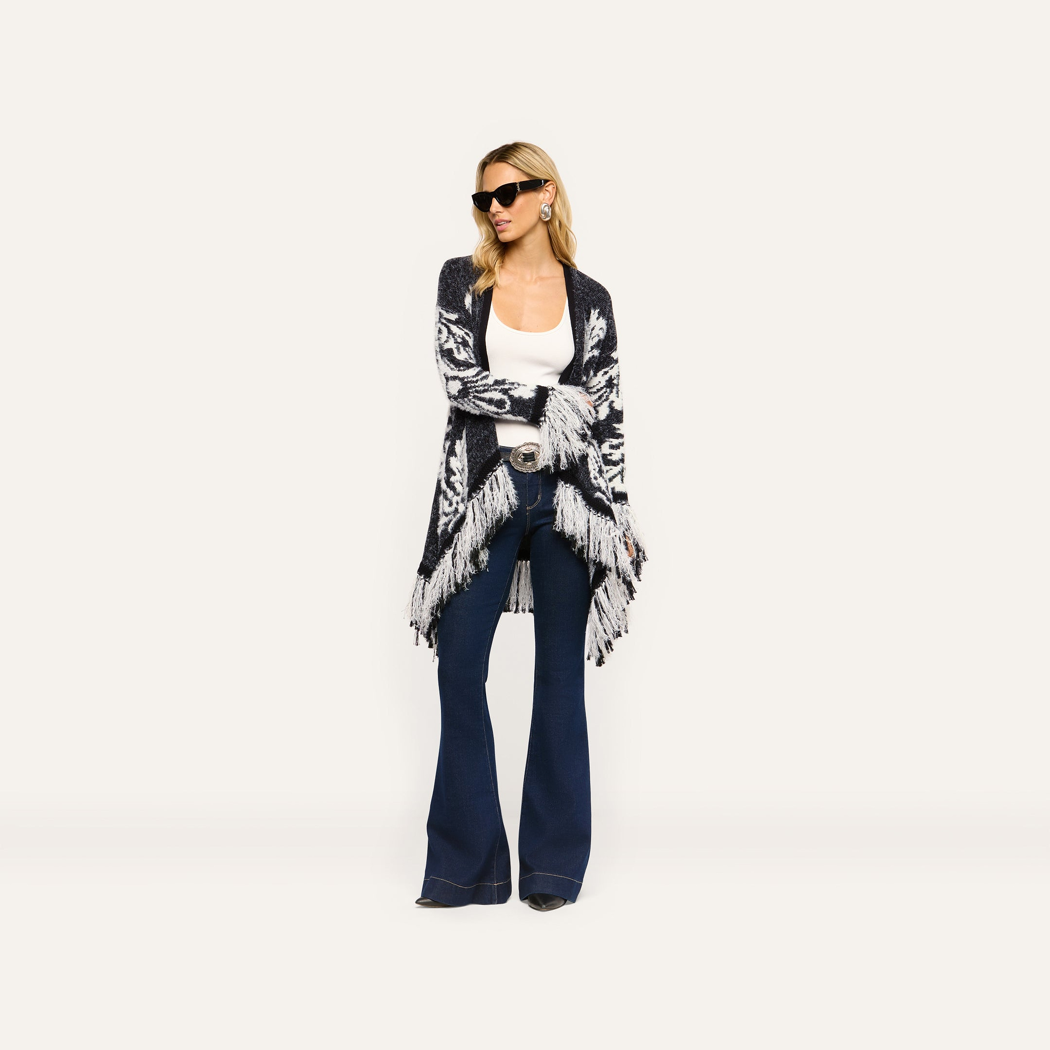 A woman wearing a black and white patterned Naiad Jacquard Fringe Cardigan over a white tank top and dark blue flare jeans.