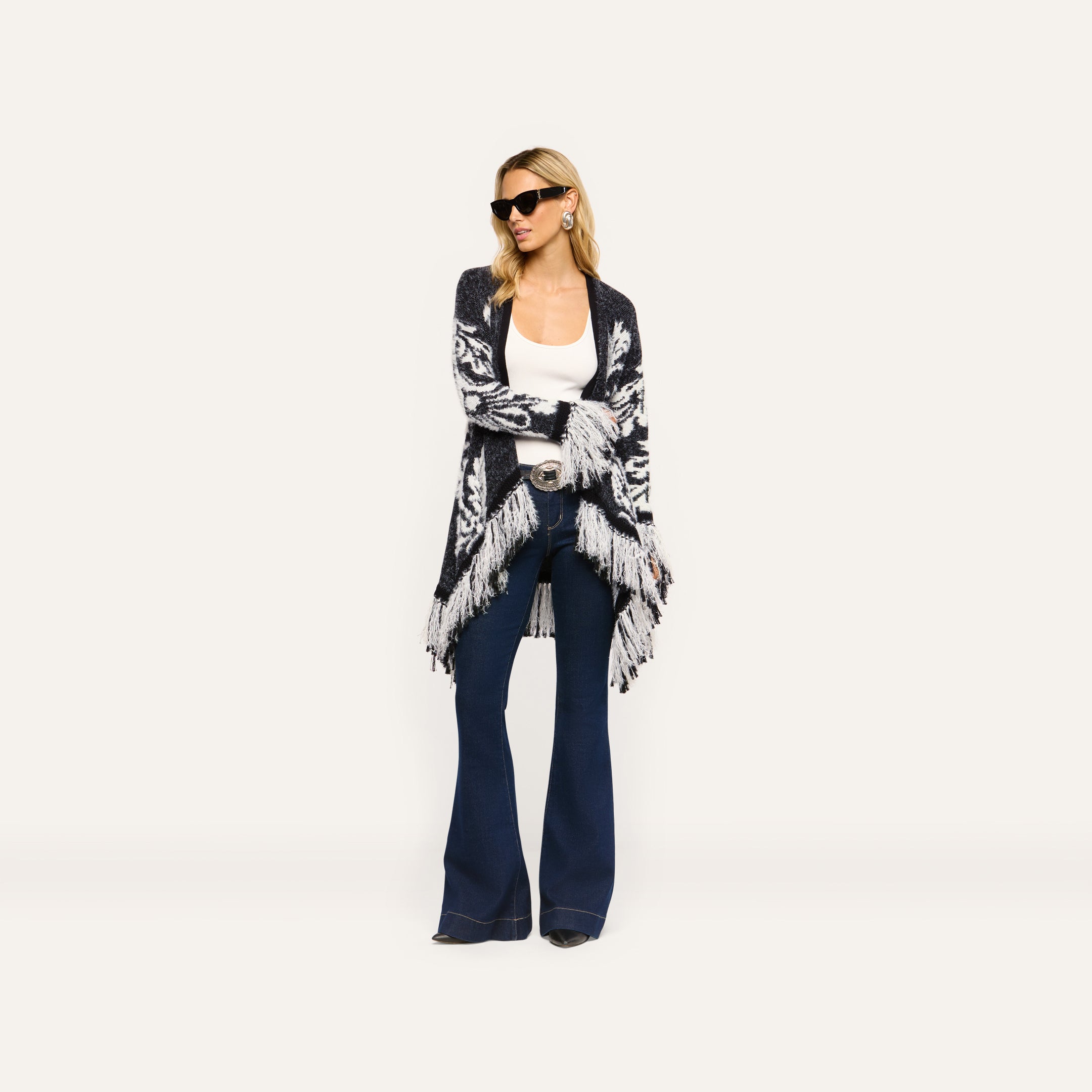 A woman wearing a black and white patterned Naiad Jacquard Fringe Cardigan over a white tank top and dark blue flare jeans.