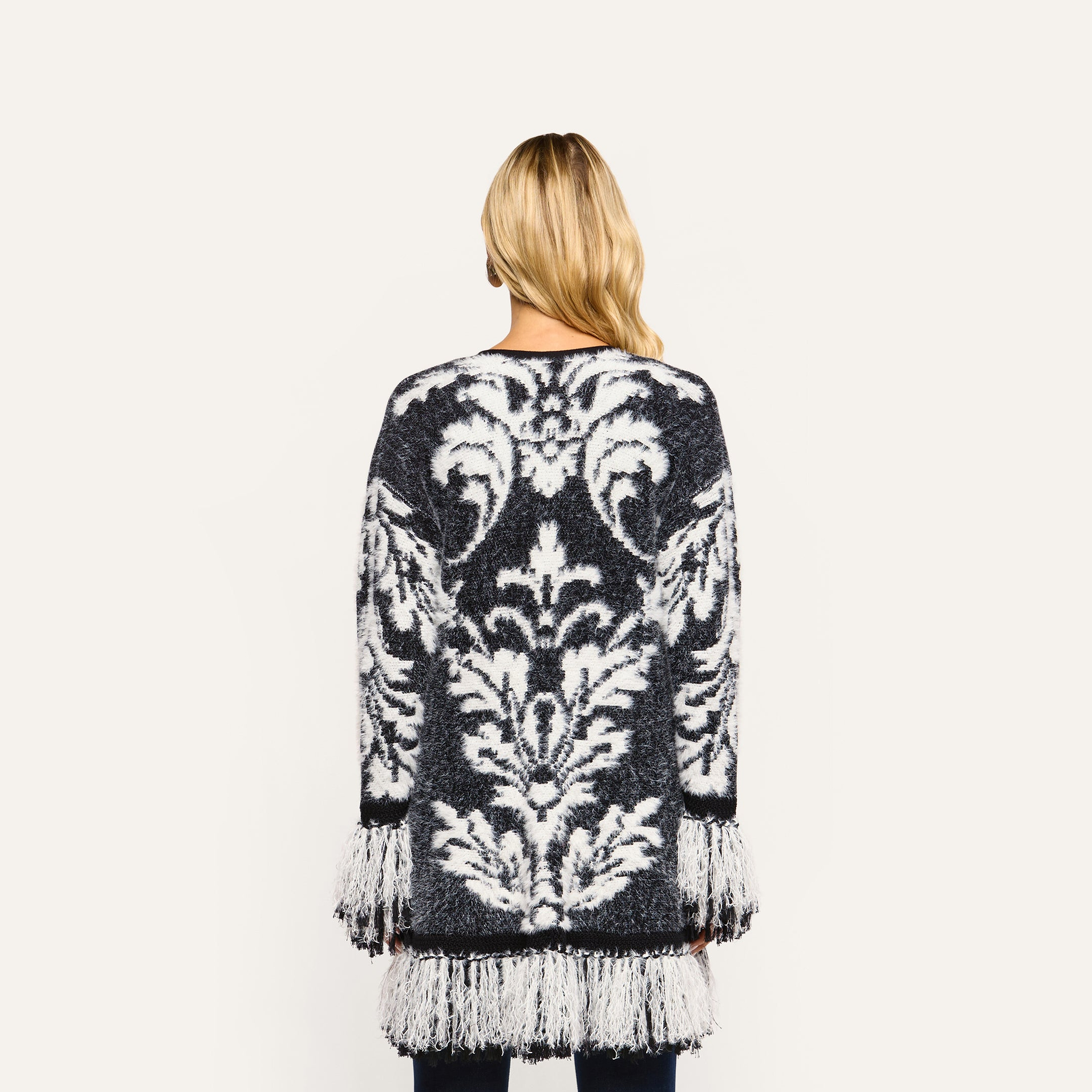 Back view of a person wearing a black and white Naiad Jacquard Fringe Cardigan, a relaxed-fit style featuring long fringe trim on the sleeves and hem.