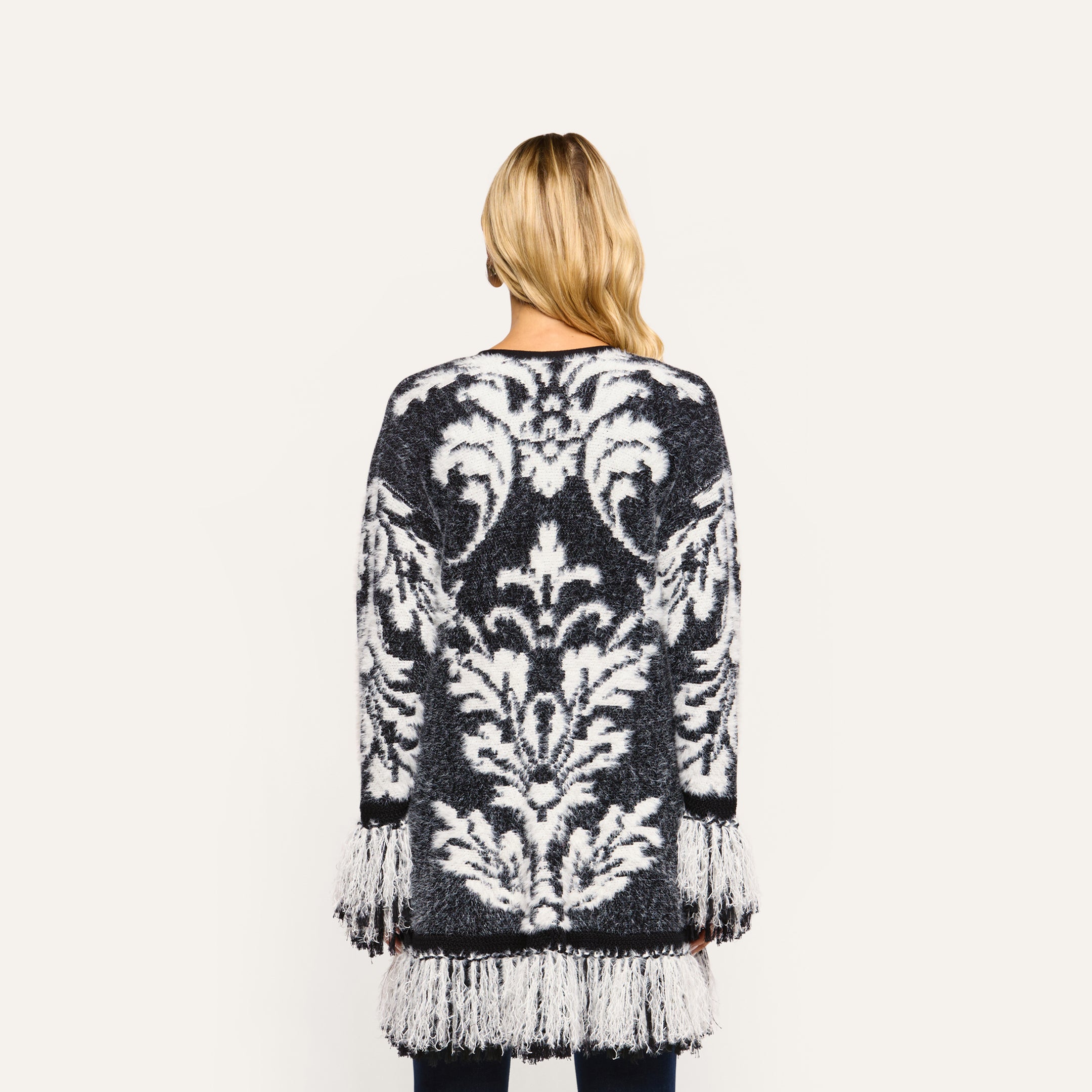 Back view of a person wearing a black and white Naiad Jacquard Fringe Cardigan, a relaxed-fit style featuring long fringe trim on the sleeves and hem.