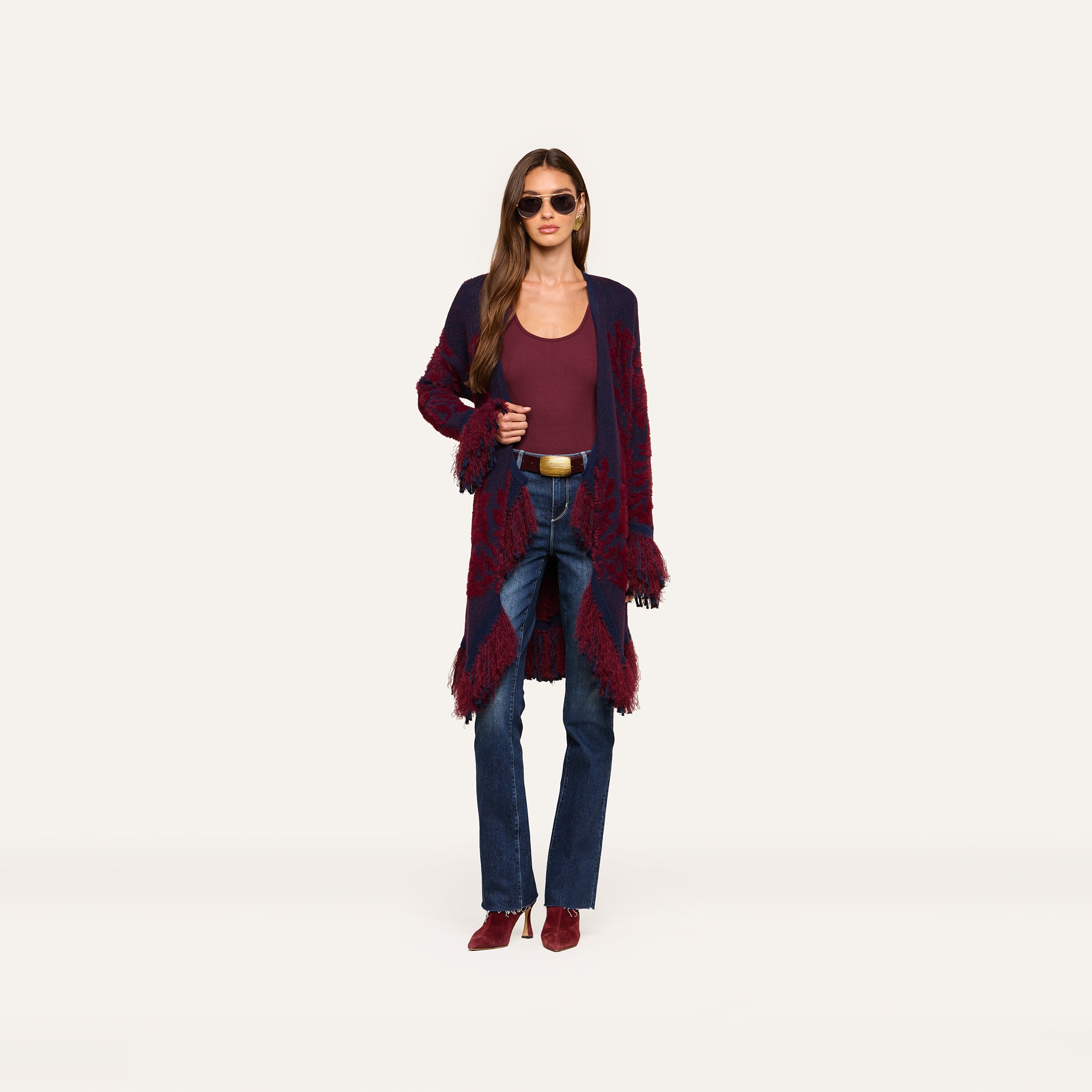 A person wearing sunglasses, a maroon top, blue jeans, maroon shoes, and the Naiad Jacquard Fringe Cardigan stands against a plain white background.