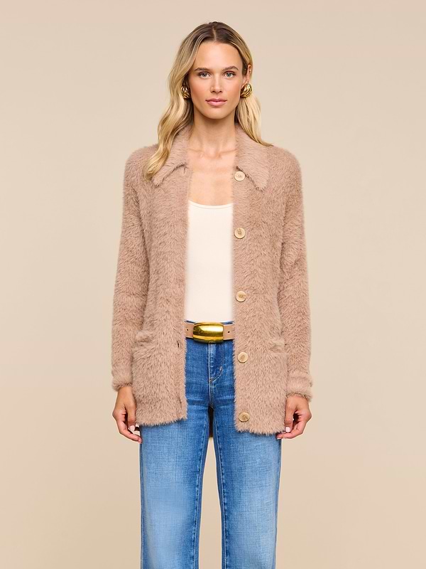 A woman with wavy blonde hair wears the Mist Cardigan—an oversized beige fuzzy piece crafted from ultra-soft yarn—layered over a cream top and blue jeans, standing against a plain light background.