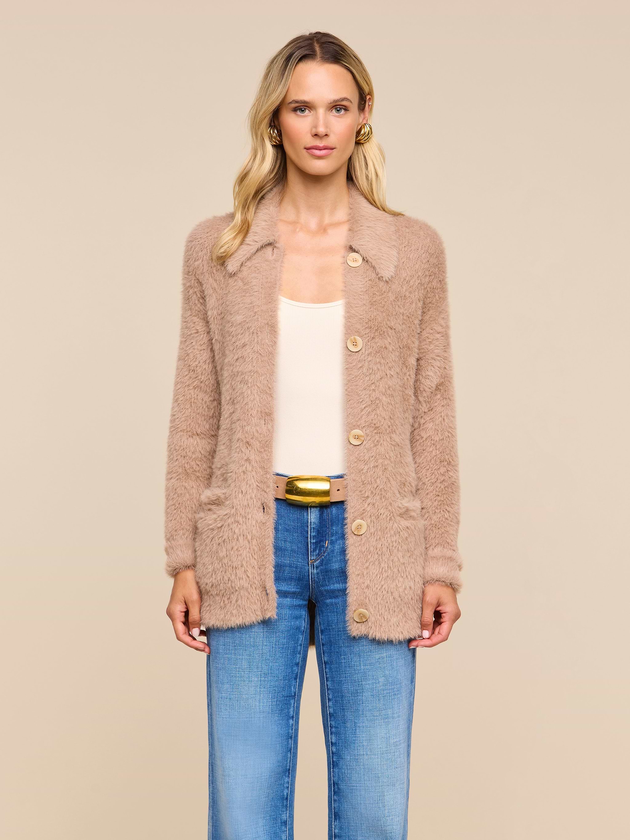 A woman with wavy blonde hair wears the Mist Cardigan—an oversized beige fuzzy piece crafted from ultra-soft yarn—layered over a cream top and blue jeans, standing against a plain light background.