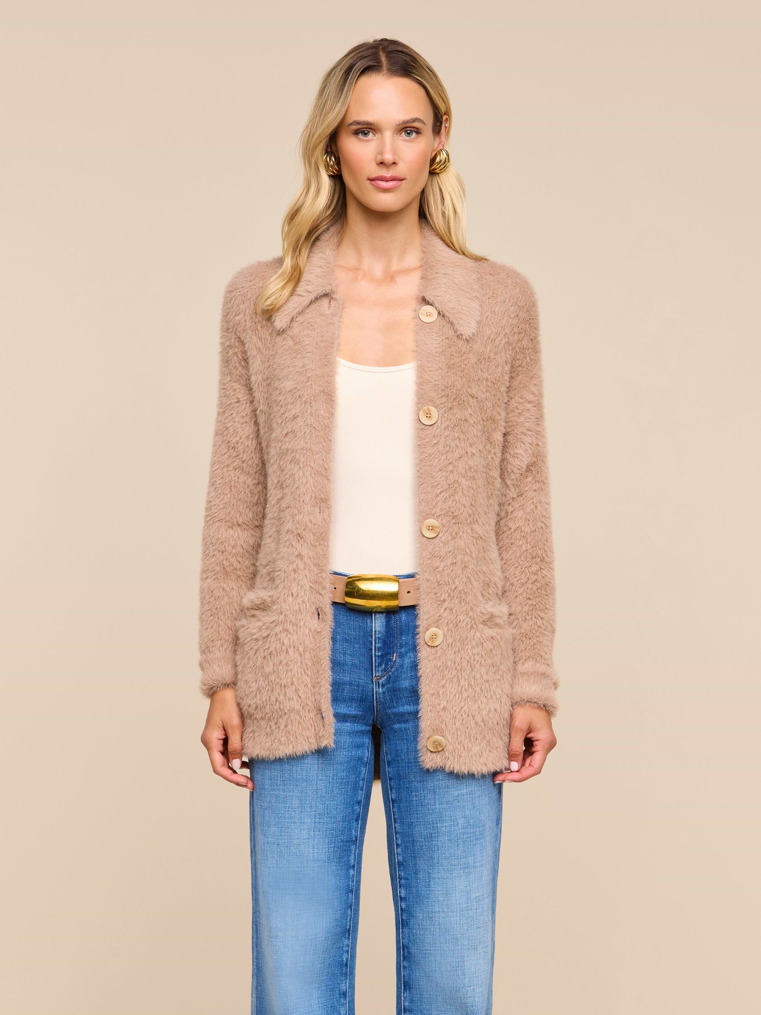A woman with wavy blonde hair wears the Mist Cardigan—an oversized beige fuzzy piece crafted from ultra-soft yarn—layered over a cream top and blue jeans, standing against a plain light background.
