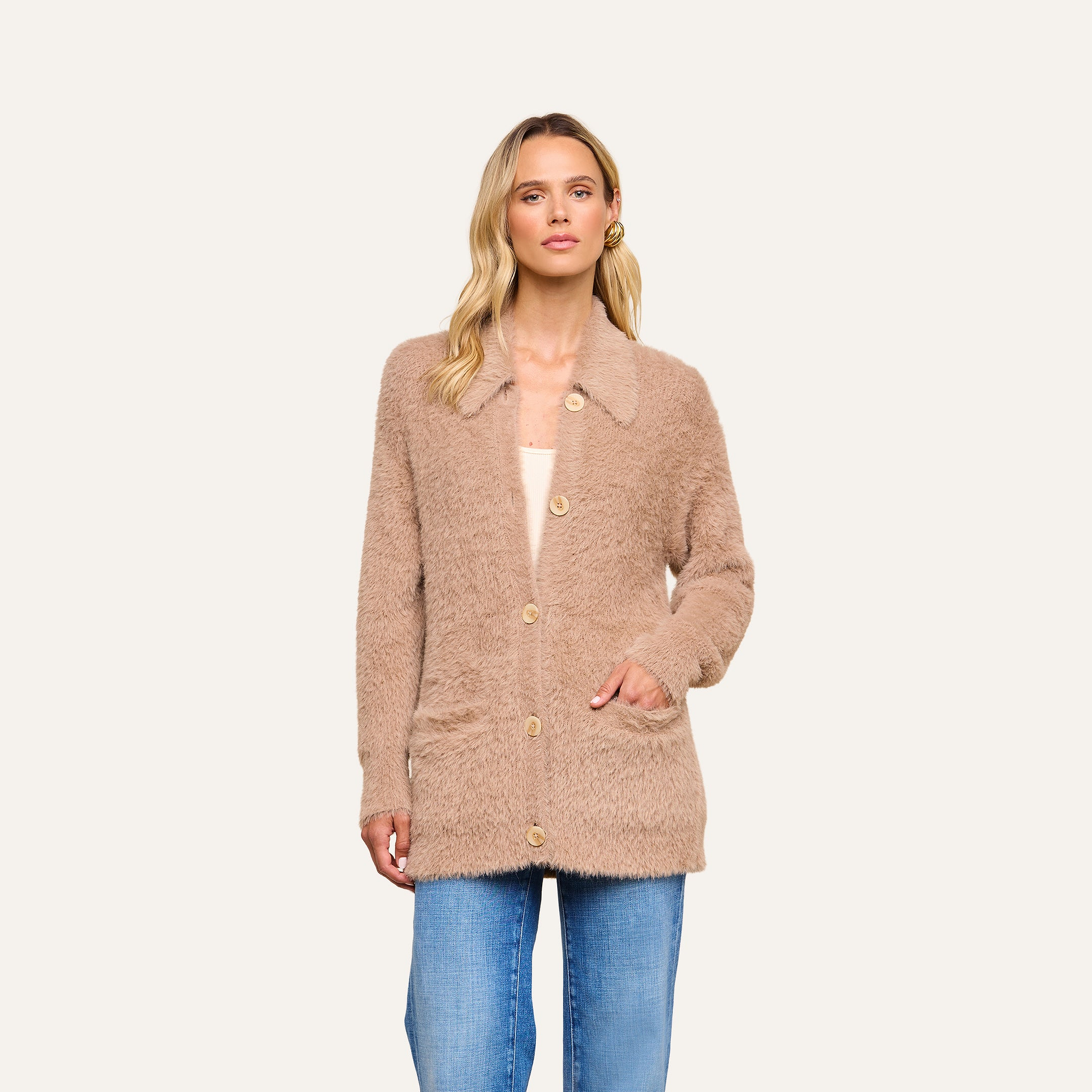 A woman wearing the Mist Cardigan—an oversized, fuzzy light brown button-up crafted from ultra-soft yarn—over a cream top and blue jeans stands against a plain light background.