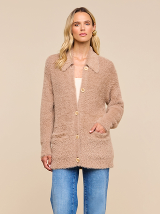 A woman wearing the Mist Cardigan—a cozy, light brown button-up with front pockets—paired with light blue jeans stands against a beige background.