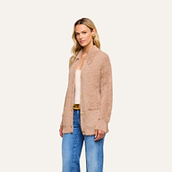 A woman with long blonde hair wears the Mist Cardigan—an oversized beige fuzzy cardigan crafted from ultra-soft yarn—over a white top and blue jeans, standing against a plain light background.