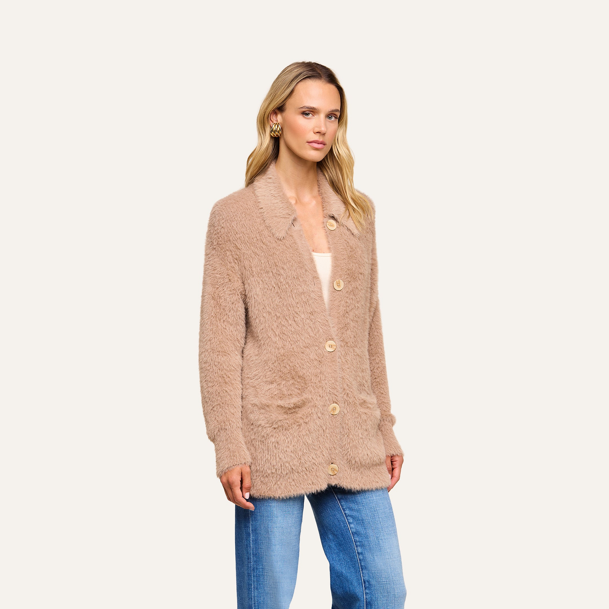 A woman stands against a plain light background wearing the Mist Cardigan—an ultra-soft, beige fuzzy cardigan with buttons—layered over a light top and paired with blue jeans.