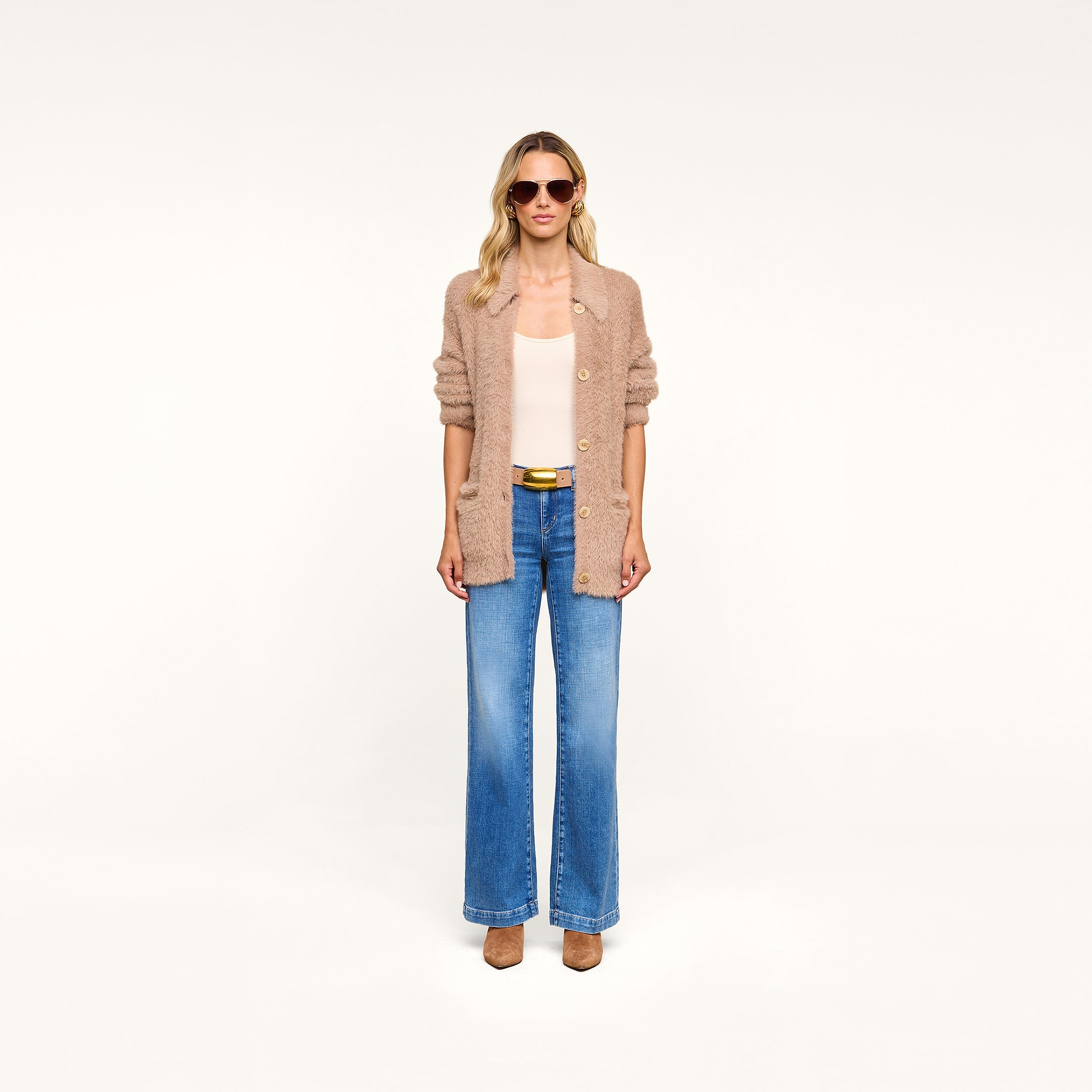 Wearing sunglasses, a white top, blue wide-leg jeans, tan shoes, and the Mist Cardigan—an oversized beige cardigan crafted from ultra-soft yarn—a person stands against a plain white background.