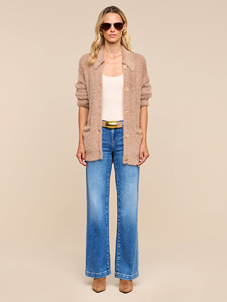 Woman wearing the Mist Cardigan in beige, styled with a cream top, wide-leg blue jeans, a brown belt, and brown boots. She stands against a plain beige background with sunglasses on.
