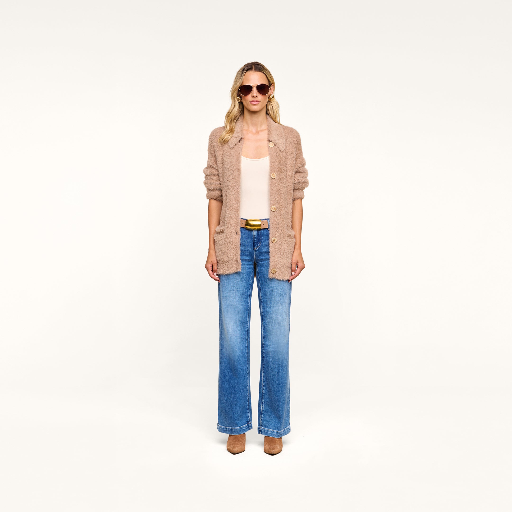Wearing sunglasses, a white top, blue wide-leg jeans, tan shoes, and the Mist Cardigan—an oversized beige cardigan crafted from ultra-soft yarn—a person stands against a plain white background.