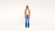Woman wearing sunglasses, a tan cardigan over a light top, wide-leg blue jeans, and brown shoes stands against a plain white background.