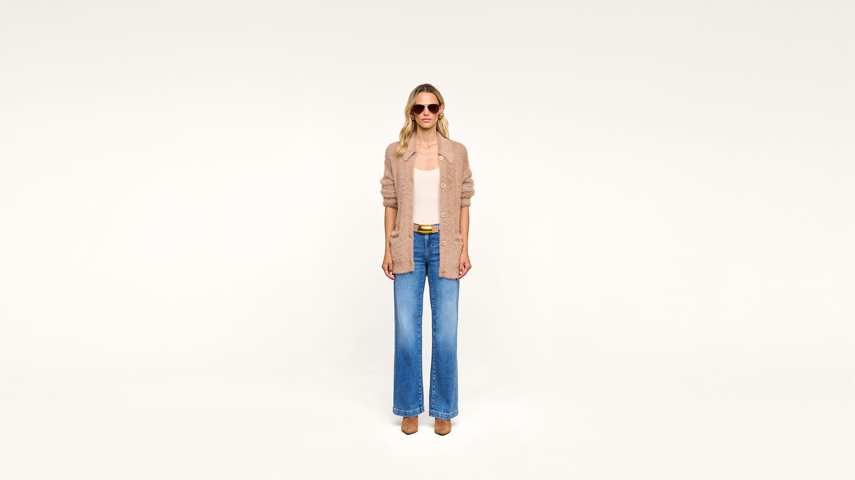 Woman wearing sunglasses, a tan cardigan over a light top, wide-leg blue jeans, and brown shoes stands against a plain white background.
