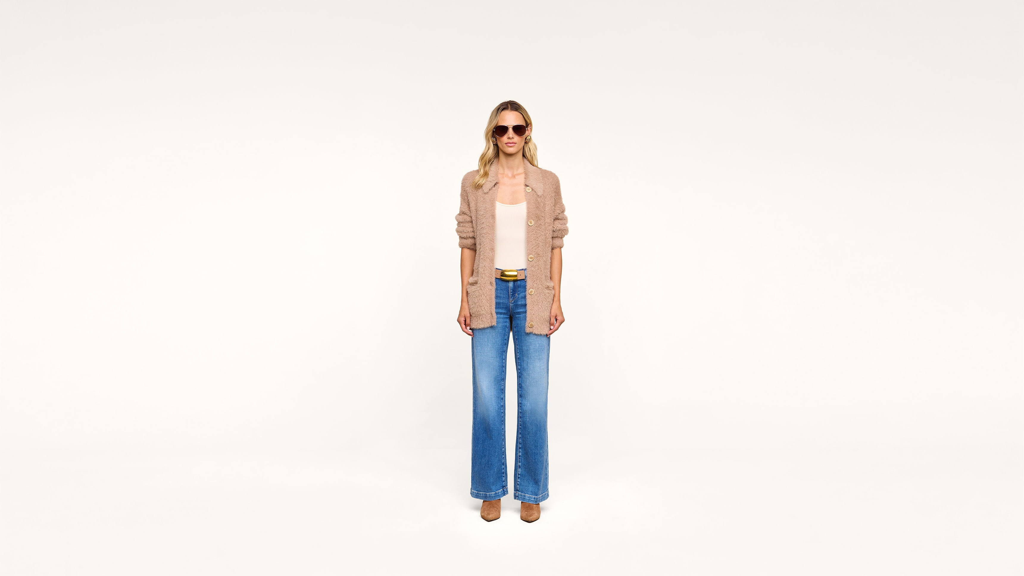 Woman wearing sunglasses, a tan cardigan over a light top, wide-leg blue jeans, and brown shoes stands against a plain white background.