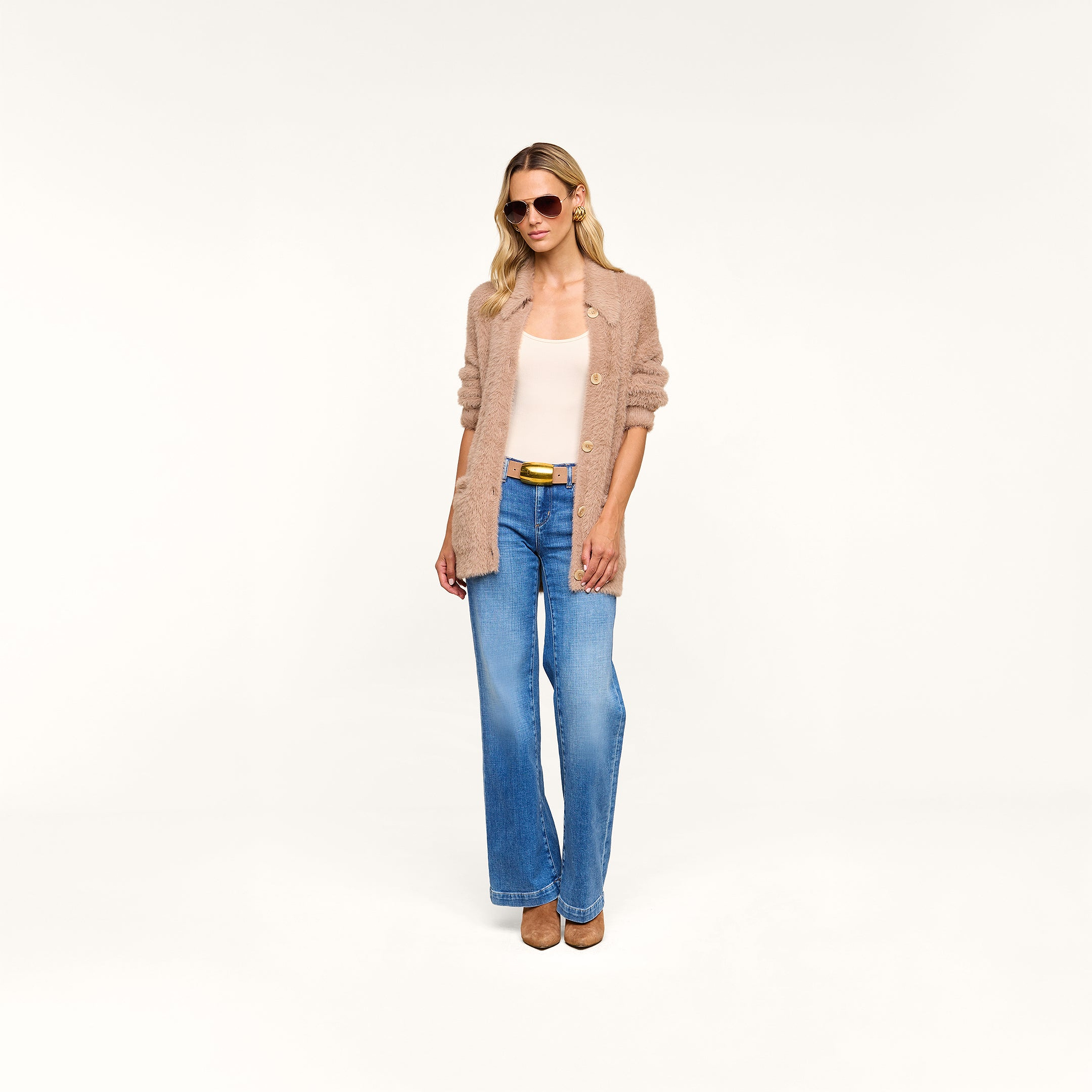 Wearing sunglasses and beige heels, a woman stands against a plain white background dressed in the Mist Cardigan—an oversized beige layering piece crafted from ultra-soft yarn—paired with a white top and blue jeans.
