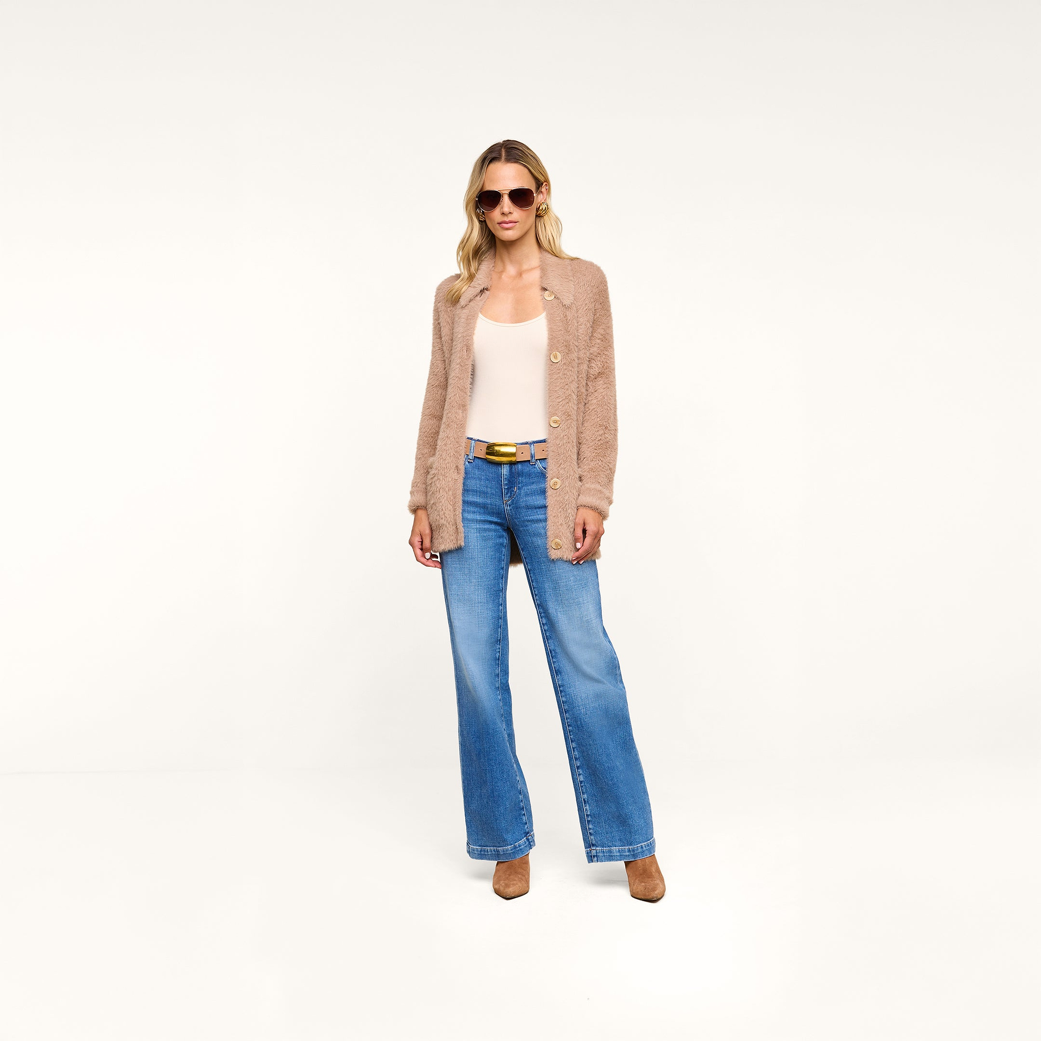 Wearing the Mist Cardigan, a person styled in sunglasses, a white top, blue wide-leg jeans, and brown heels stands in a minimal, white studio setting.