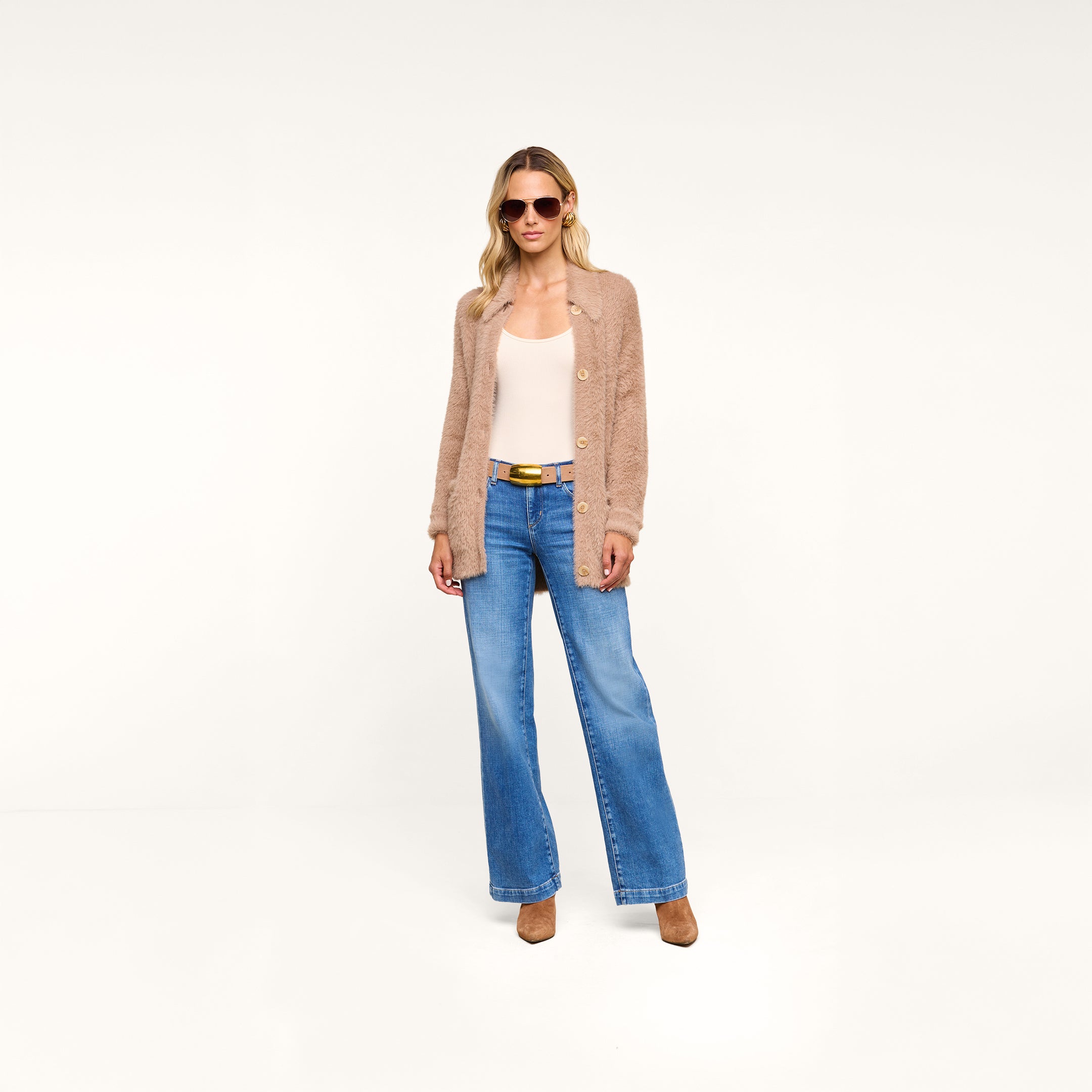 Wearing the Mist Cardigan, a person styled in sunglasses, a white top, blue wide-leg jeans, and brown heels stands in a minimal, white studio setting.