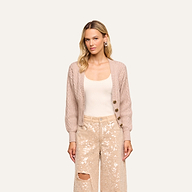 A woman with long blonde hair wears the Hail Cabled Cardigan with blouson sleeves in light taupe over a cream top and beige sequin pants featuring a rip on one knee, standing against a plain background.