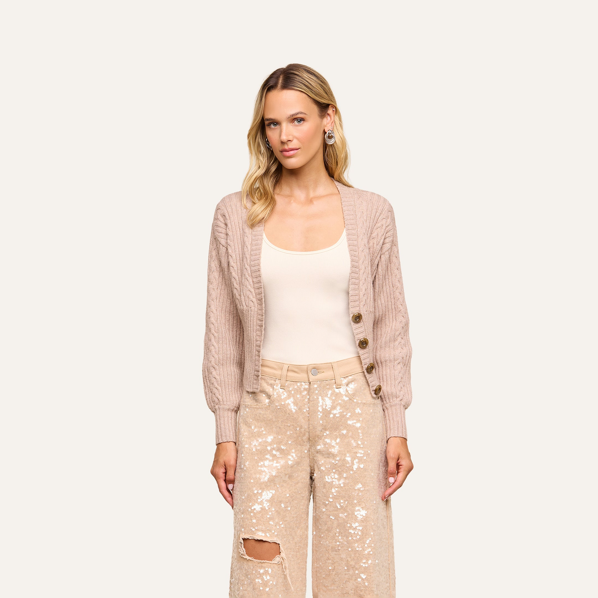 A woman with long blonde hair wears the Hail Cabled Cardigan with blouson sleeves in light taupe over a cream top and beige sequin pants featuring a rip on one knee, standing against a plain background.