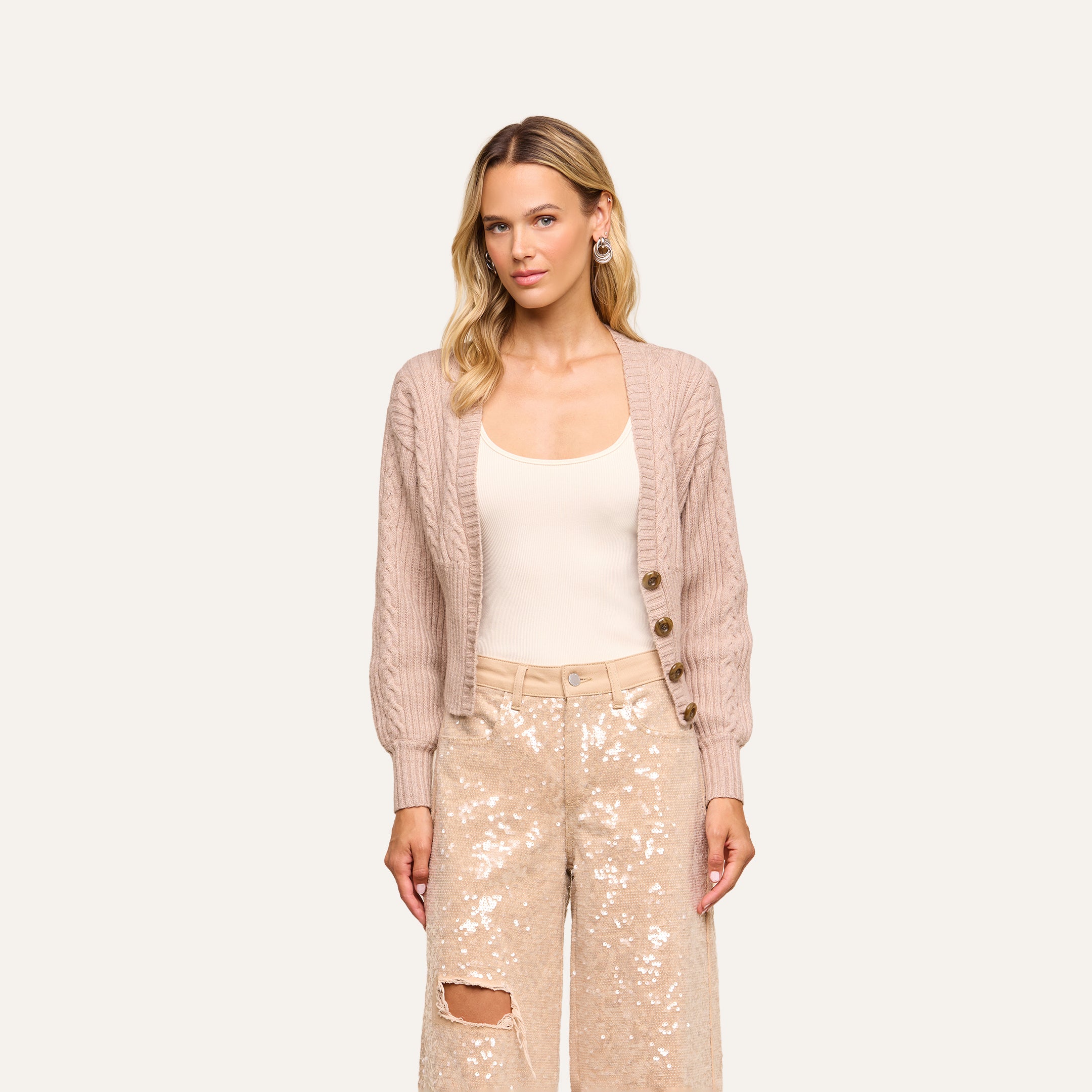 A woman with long blonde hair wears the Hail Cabled Cardigan with blouson sleeves in light taupe over a cream top and beige sequin pants featuring a rip on one knee, standing against a plain background.