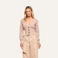 Woman with long blonde hair wearing the Hail Cabled Cardigan—a beige, deep V-neck cable knit design with blouson sleeves and large buttons—paired with light-colored, high-waisted sequined pants, standing against a plain light background.