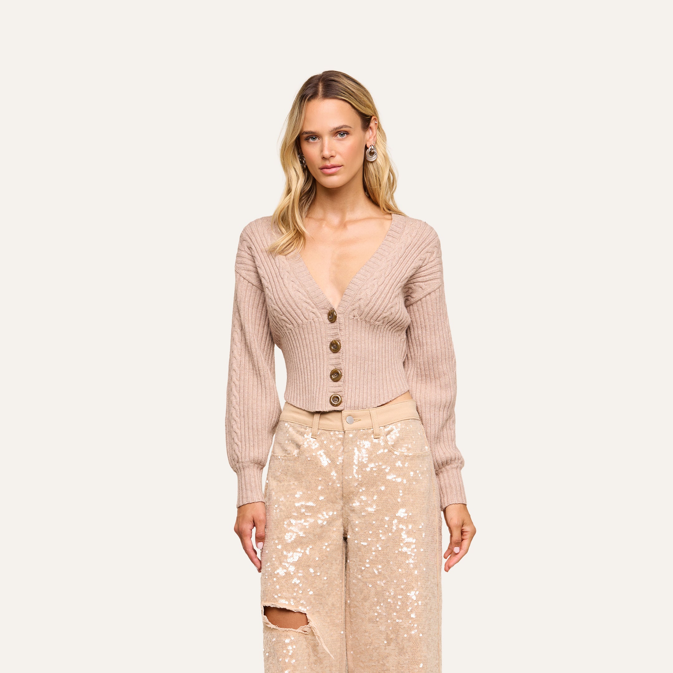 Woman with long blonde hair wearing the Hail Cabled Cardigan—a beige, deep V-neck cable knit design with blouson sleeves and large buttons—paired with light-colored, high-waisted sequined pants, standing against a plain light background.