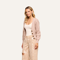A woman with long blonde hair wears the Hail Cabled Cardigan, featuring blouson sleeves, over a white tank top and light-colored sequined pants. She stands against an off-white background, looking toward the camera with a neutral expression.