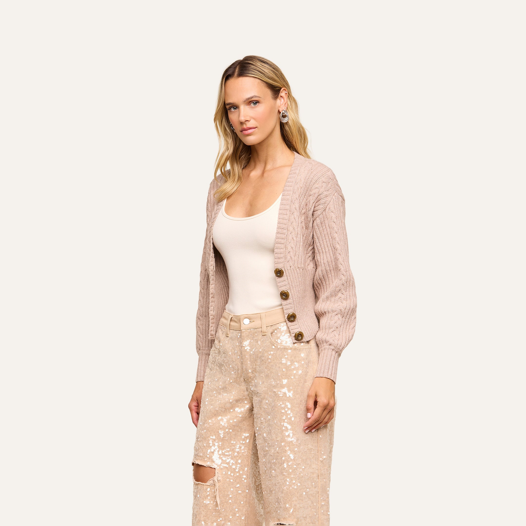 A woman with long blonde hair wears the Hail Cabled Cardigan, featuring blouson sleeves, over a white tank top and light-colored sequined pants. She stands against an off-white background, looking toward the camera with a neutral expression.