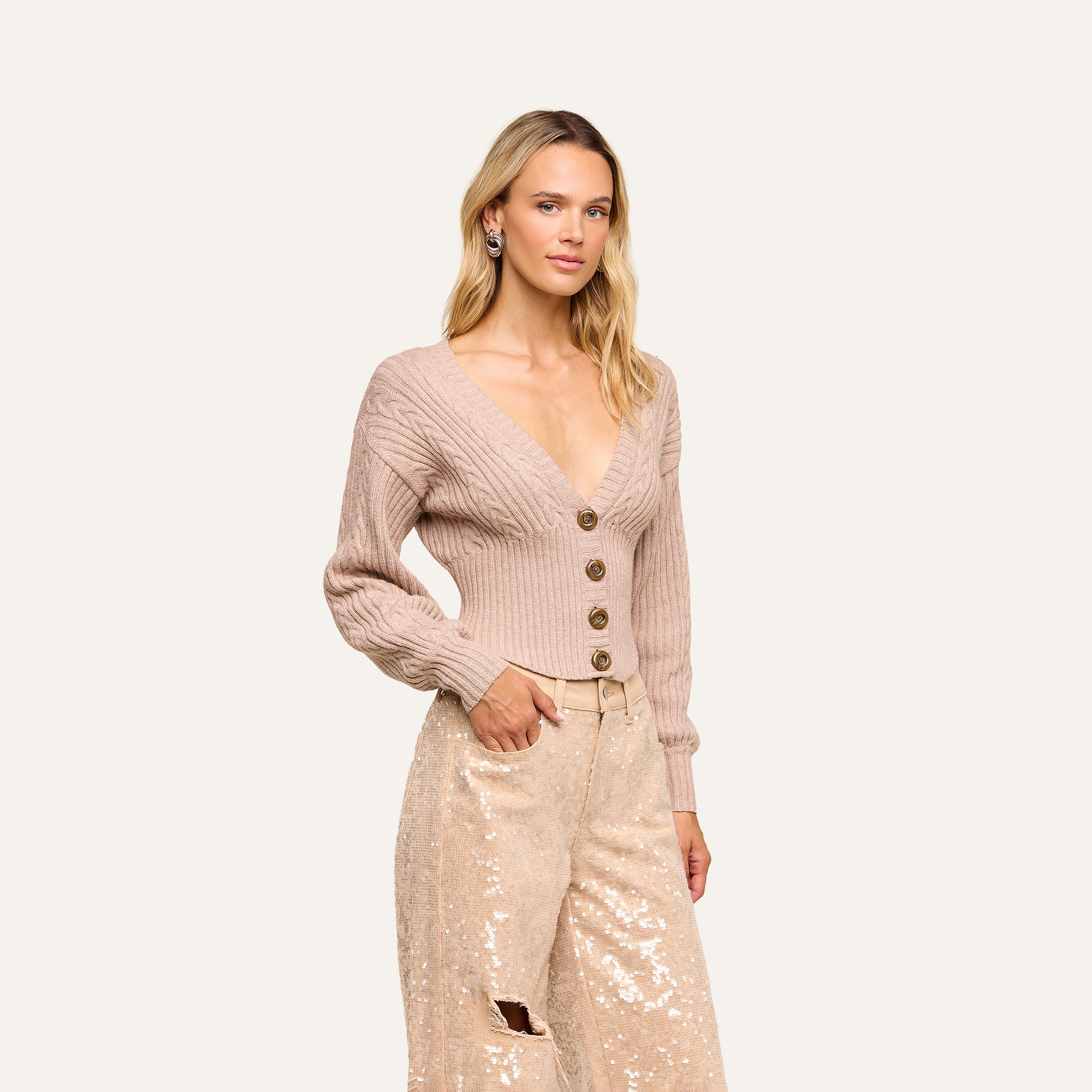 A woman poses with one hand in her pocket against a plain background, wearing the Hail Cabled Cardigan—a light pink ribbed cropped style featuring blouson sleeves and large buttons—paired with cream sequin pants.