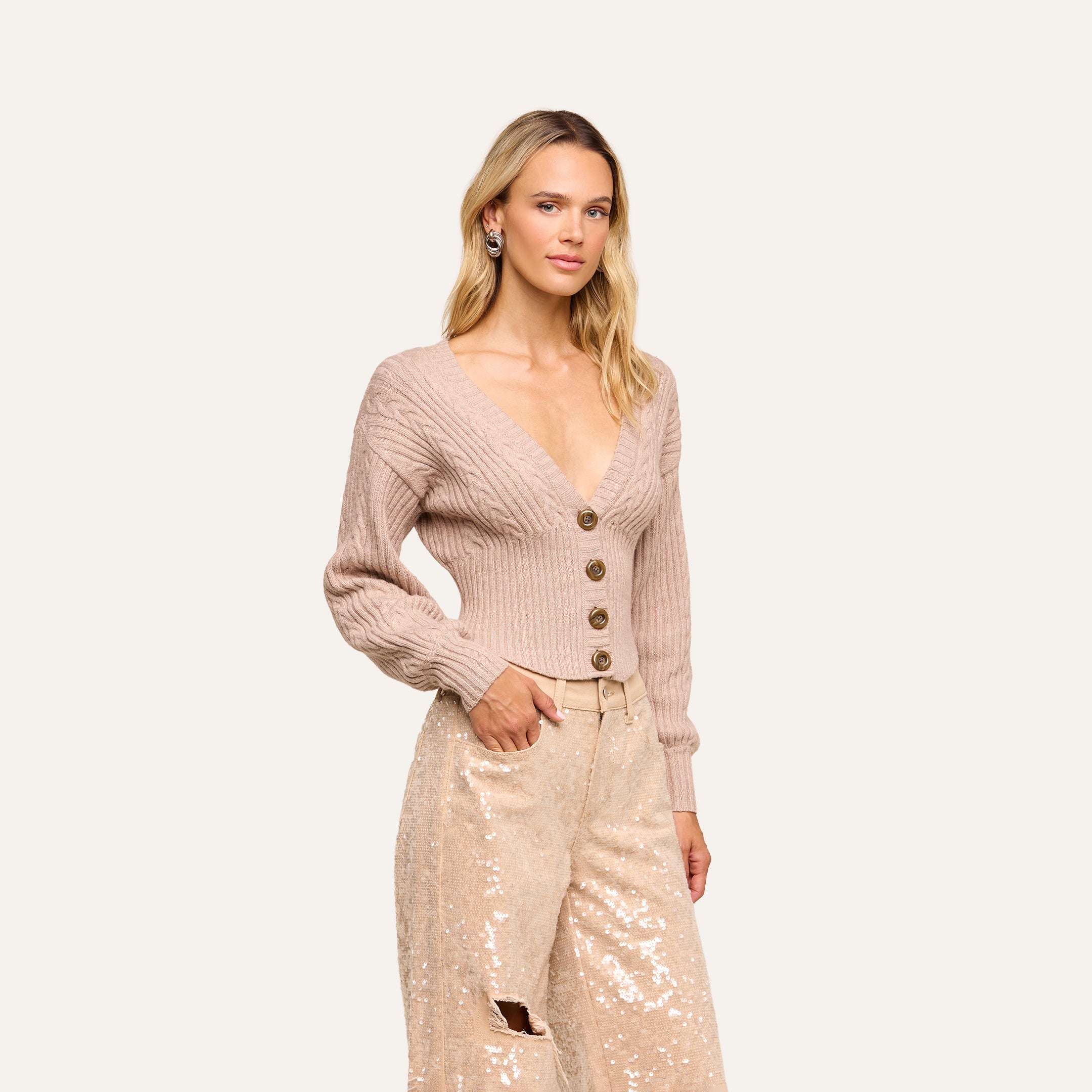 A woman poses with one hand in her pocket against a plain background, wearing the Hail Cabled Cardigan—a light pink ribbed cropped style featuring blouson sleeves and large buttons—paired with cream sequin pants.