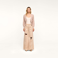 A woman stands facing forward against a plain white background, wearing the Hail Cabled Cardigan with blouson sleeves, paired with a cream tank top and wide-legged, sequined, ripped beige pants and heels.