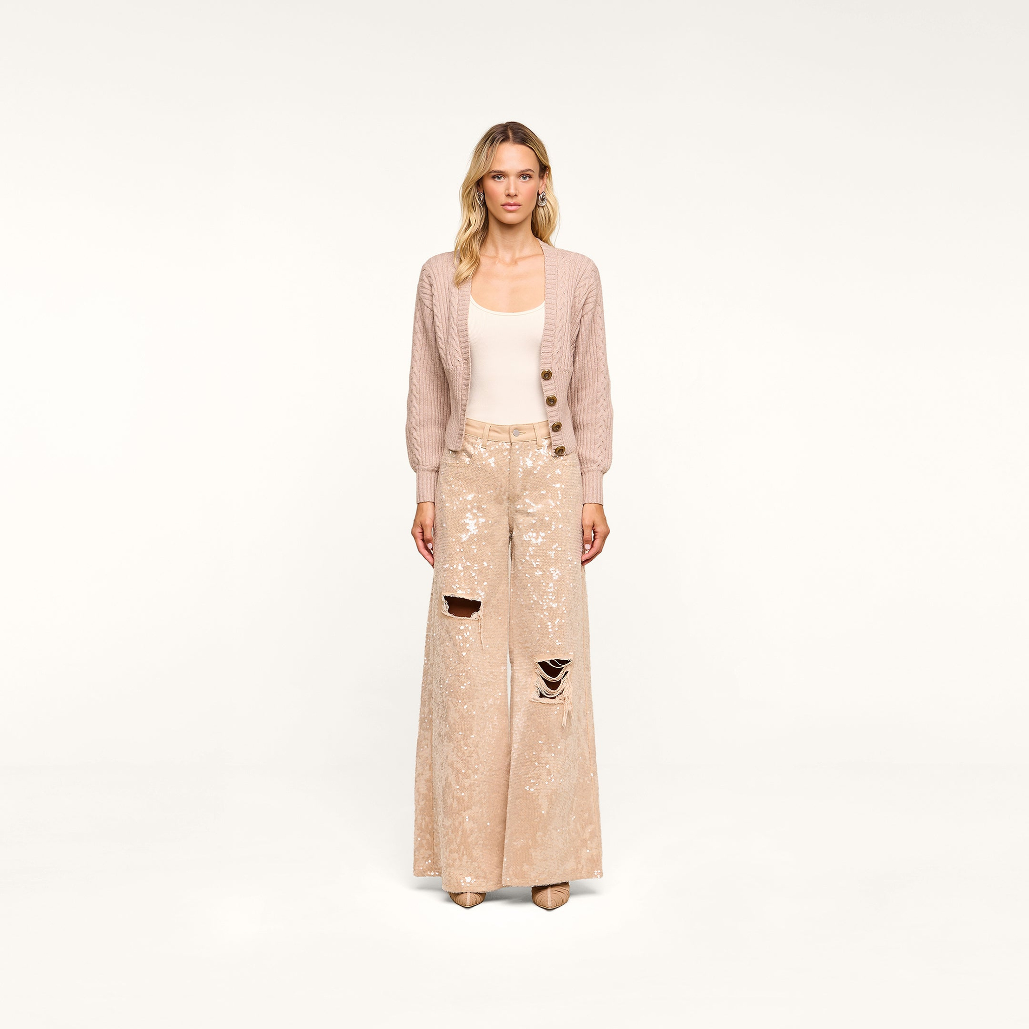 A woman stands facing forward against a plain white background, wearing the Hail Cabled Cardigan with blouson sleeves, paired with a cream tank top and wide-legged, sequined, ripped beige pants and heels.