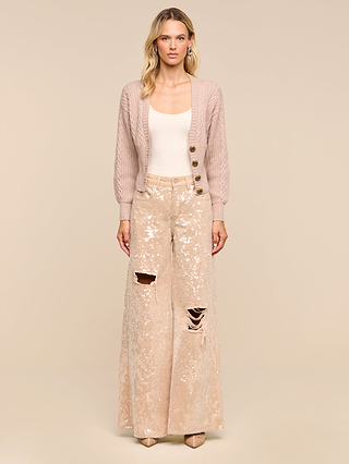 A woman stands against a beige background wearing the Hail Cabled Cardigan with blouson sleeves, paired with a cream tank top and wide-leg sequined pants featuring distressed rips on one leg. She has long, wavy blonde hair and neutral-toned heels.