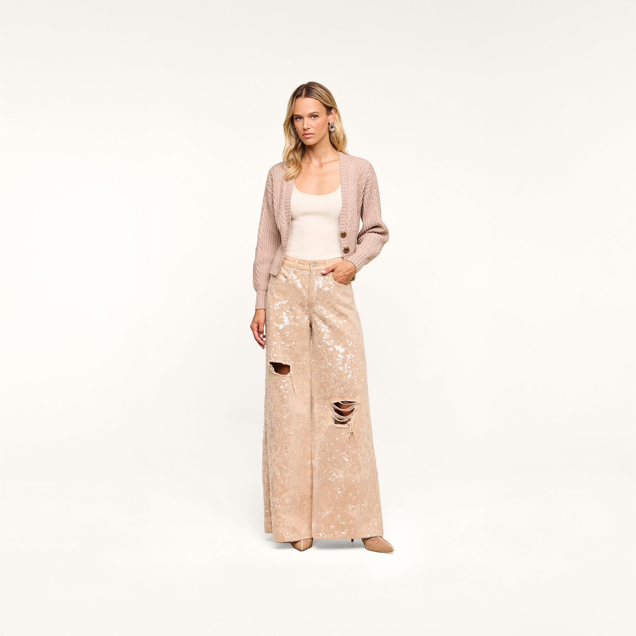 A woman stands against a plain background, wearing the Hail Cabled Cardigan in light pink with blouson sleeves, a beige top, and wide-legged sequined pants with ripped knees. She has a neutral expression and beige heels.