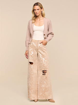 A woman wears the Hail Cabled Cardigan—a light, cropped cardigan with blouson sleeves—layered over a cream tank top and paired with wide-leg, high-waisted sequin pants featuring rips. She stands against a plain beige background.