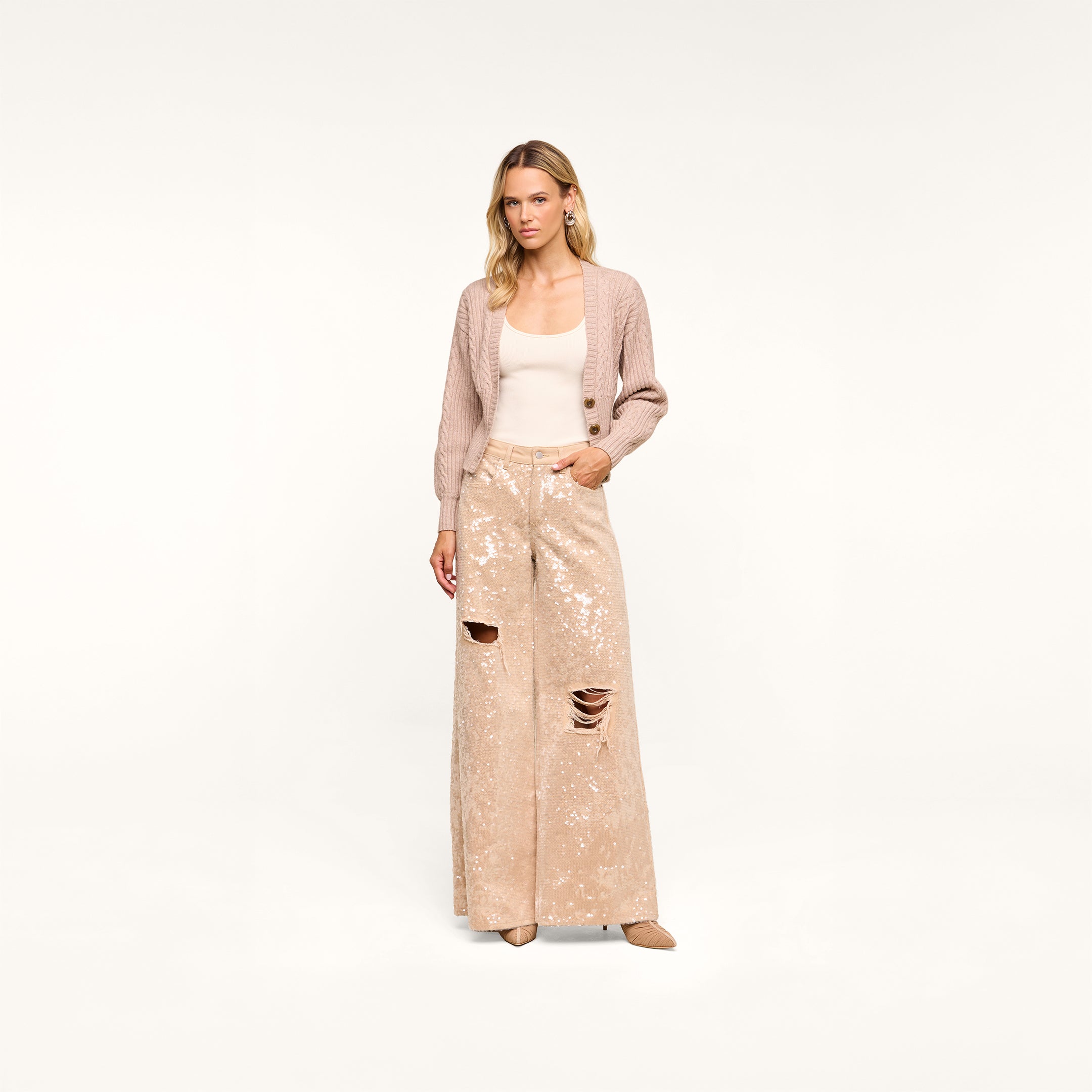 A woman stands against a plain background, wearing the Hail Cabled Cardigan in light pink with blouson sleeves, a beige top, and wide-legged sequined pants with ripped knees. She has a neutral expression and beige heels.